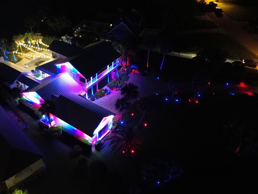 Aerial view of a home lit in purple and blue along the rooflines and pool deck